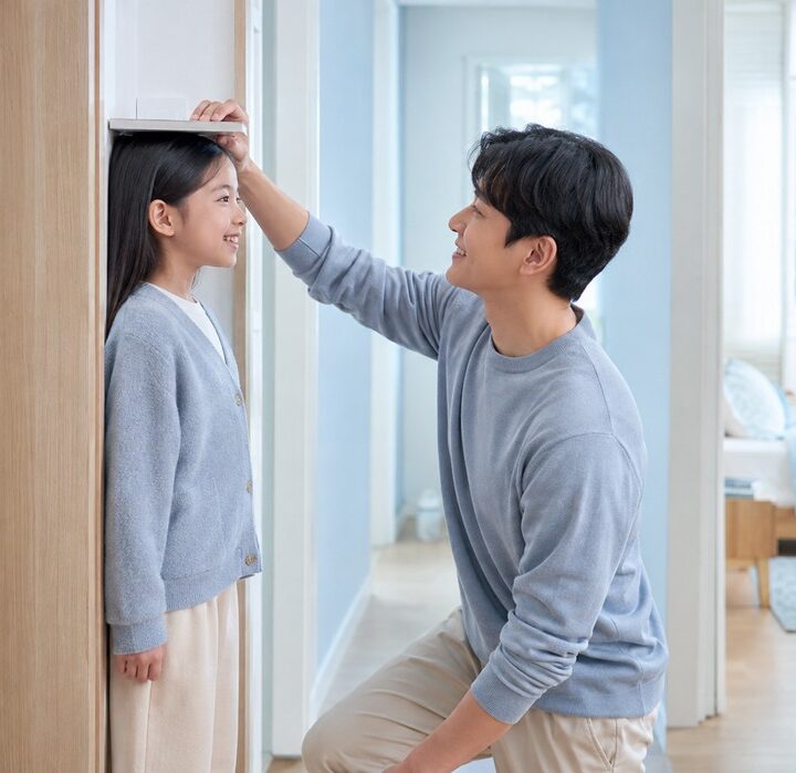 나무 문틀 옆 벽에서 회색 카디건을 입은 여자아이의 키를 회색 니트 차림 아빠가 손으로 재주는 모습