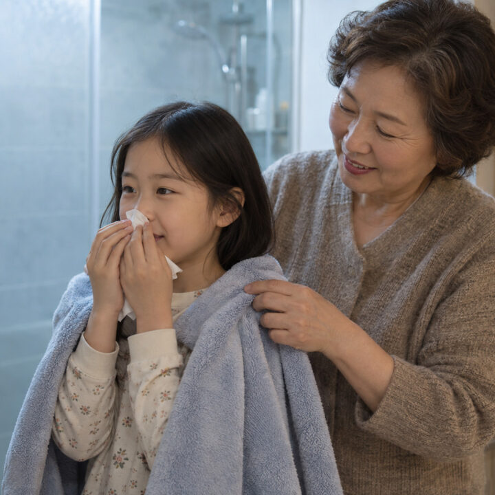 목동 부비동염 누런 콧물 – 여자아이가 코에 휴지를 대고 있고 할머니가 어깨에 수건을 둘러 주는 모습