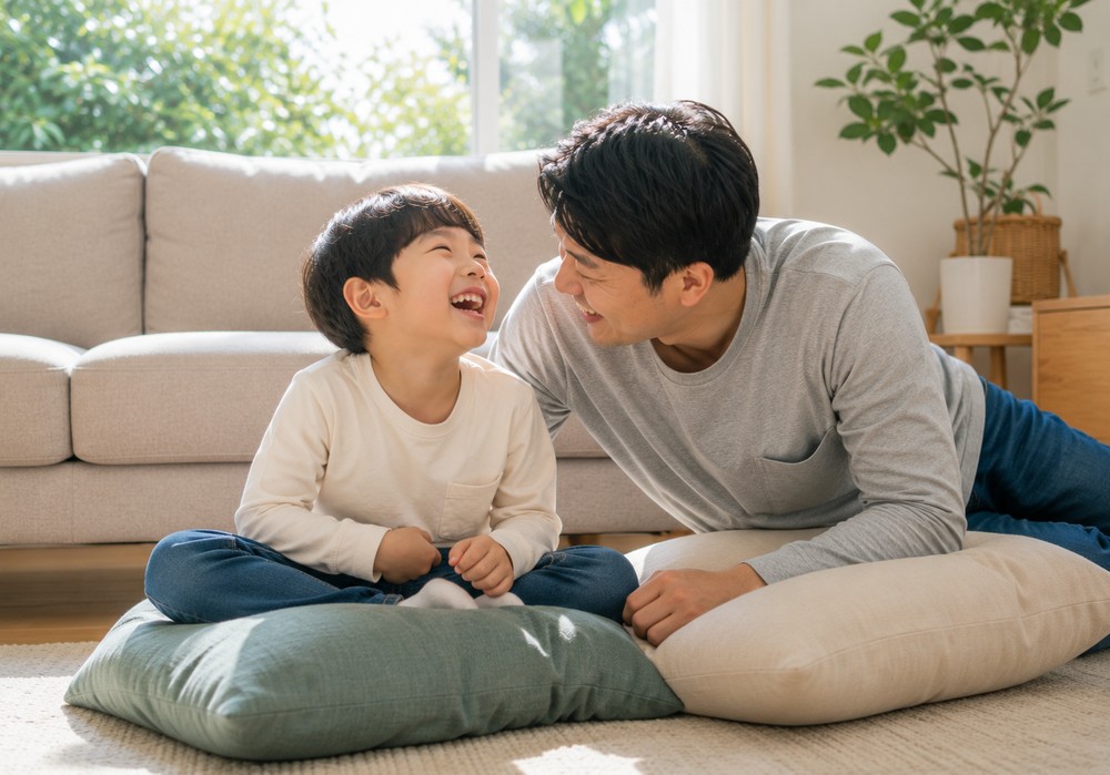 거실 바닥 쿠션 위에서 아빠와 아이가 마주보며 즐겁게 웃는 모습