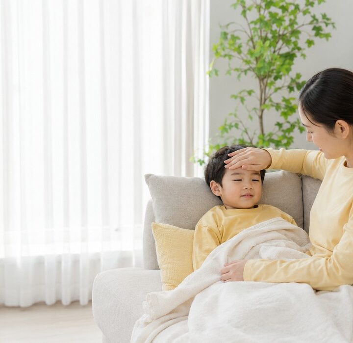 밝은 거실 소파에서 노란색 상의를 입은 남자아이가 담요를 덮고 기대어 있고, 노란 상의 엄마가 아이 이마를 짚어보는 모습