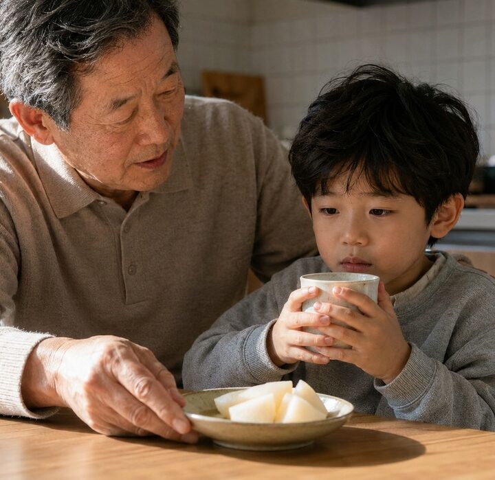 베이지 니트를 입은 할아버지가 식탁에서 배 접시를 내밀고 회색 상의 남아가 두 손으로 찻잔을 든 모습