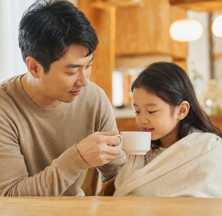 주방 식탁에서 베이지 상의 아빠가 아이보리 담요를 두른 여자아이에게 흰색 컵을 건네는 모습